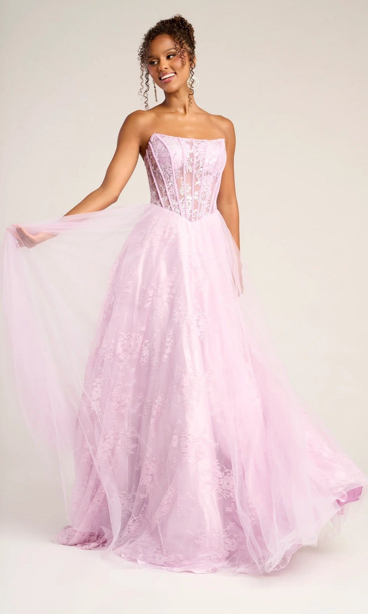 A woman stands holding out her long formal dress, the Portia and Scarlett XO023 in lilac, featuring a strapless lace ball gown design with a delicate corset bodice and a sheer tulle and lace skirt.